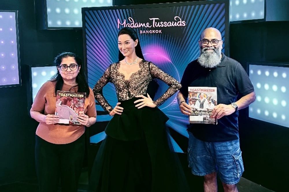 Two people stand on either side of a Madame Tussauds wax figure of a woman in a black gown, holding Toastmasters magazines, with a backdrop of illuminated panels.