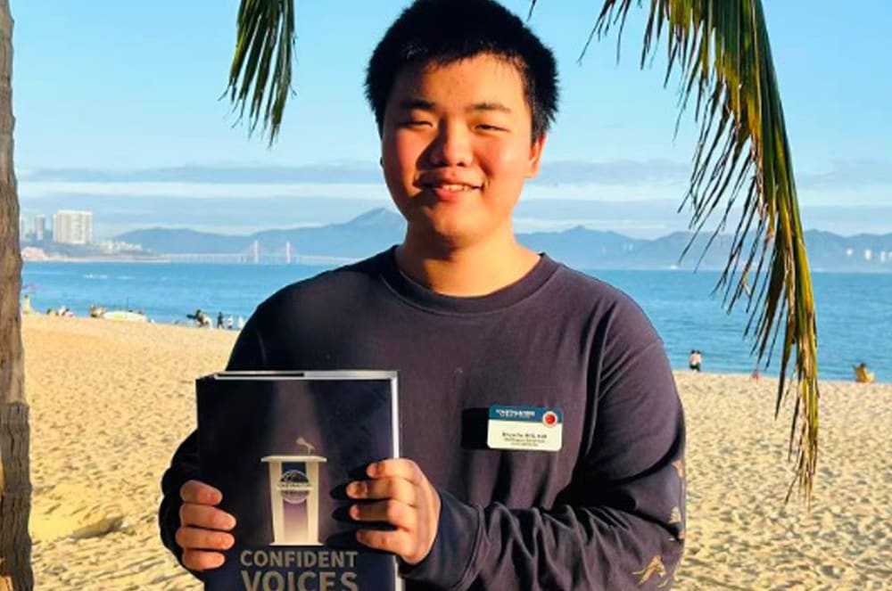 A young man holds a book on a sandy beach with palm trees and a city skyline in the background.