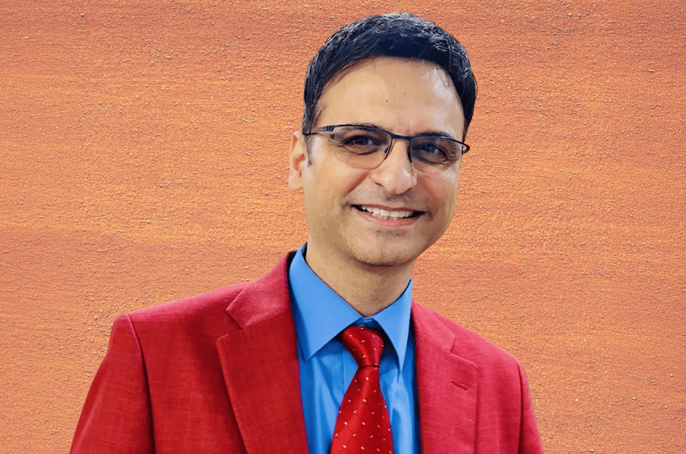 A smiling man in a red suit and blue shirt with a red polka-dot tie stands against a textured orange background.