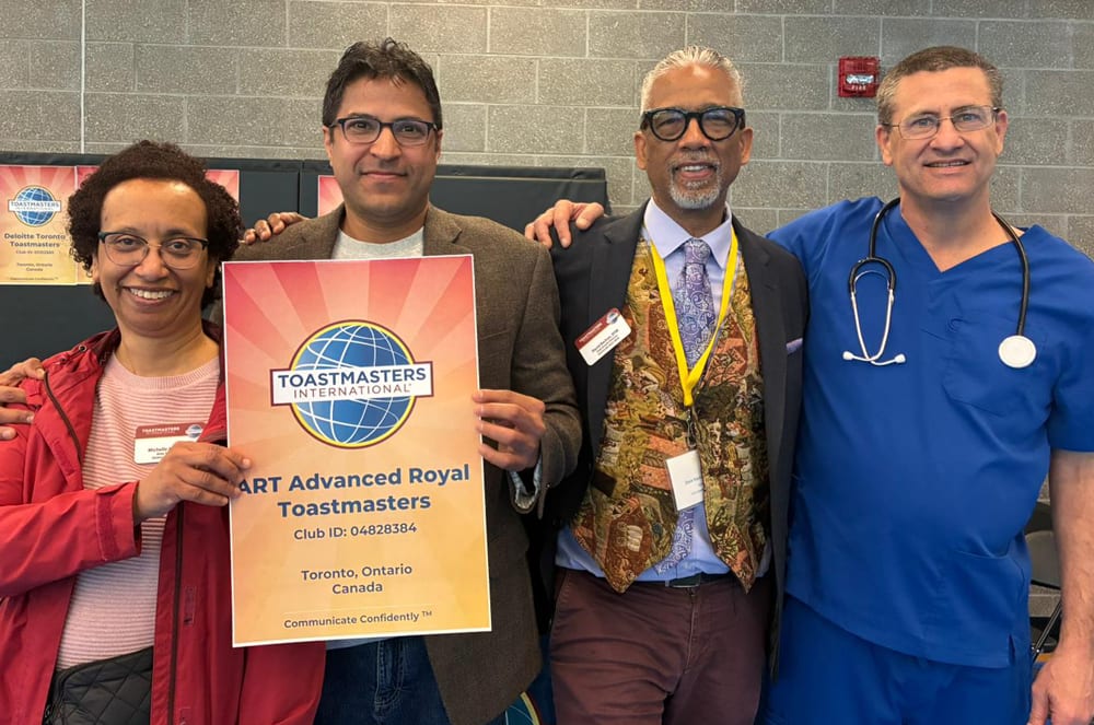 Four people stand together, holding a Toastmasters sign in the foreground, with a brick wall and a fire alarm visible in the background.