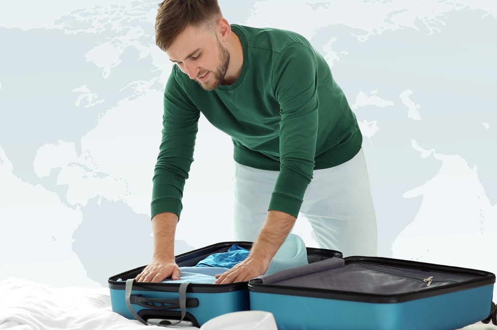 A man in a green sweater packs a blue suitcase on a white bed, with a faint world map in the background.