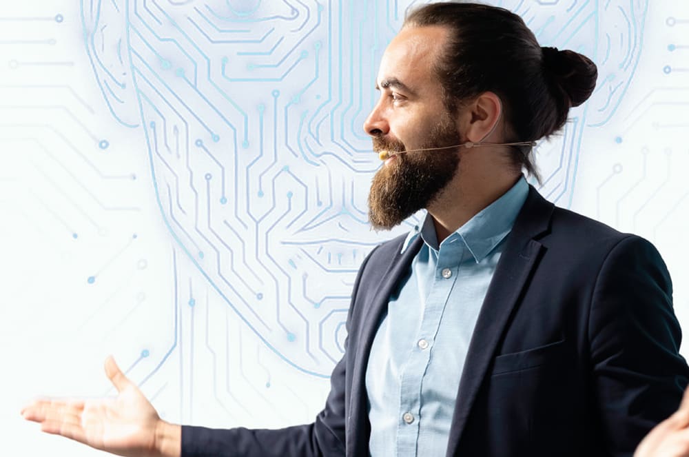 A man with a beard and bun gestures with his hands in front of a glowing, circuit-board-like outline of a human head.