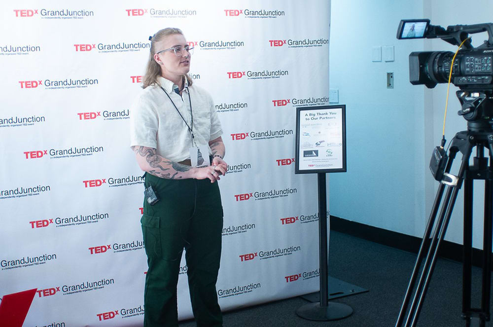 A person with tattoos on their arms stands in front of a TEDx Grand Junction backdrop, while a professional video camera on a tripod is positioned to their right.