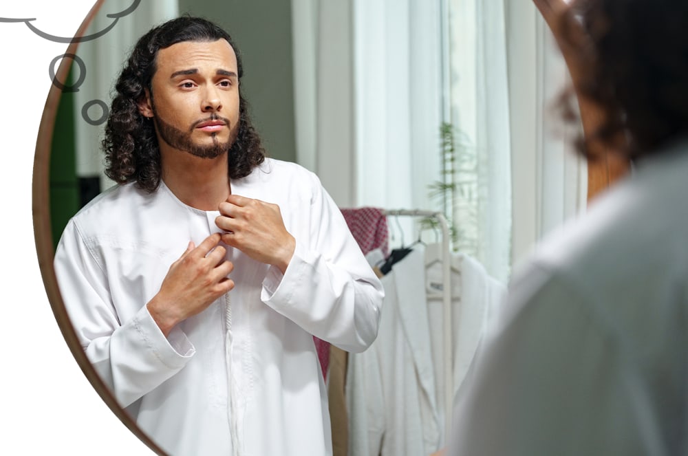 A young man with long, dark curly hair buttons up his white thobe in front of a mirror, with a blurred background of a room containing clothing on hangers and a window with sheer curtains.