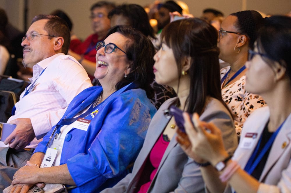 In the foreground, a woman in a blue jacket laughs heartily, while in the background, a diverse audience listens attentively.