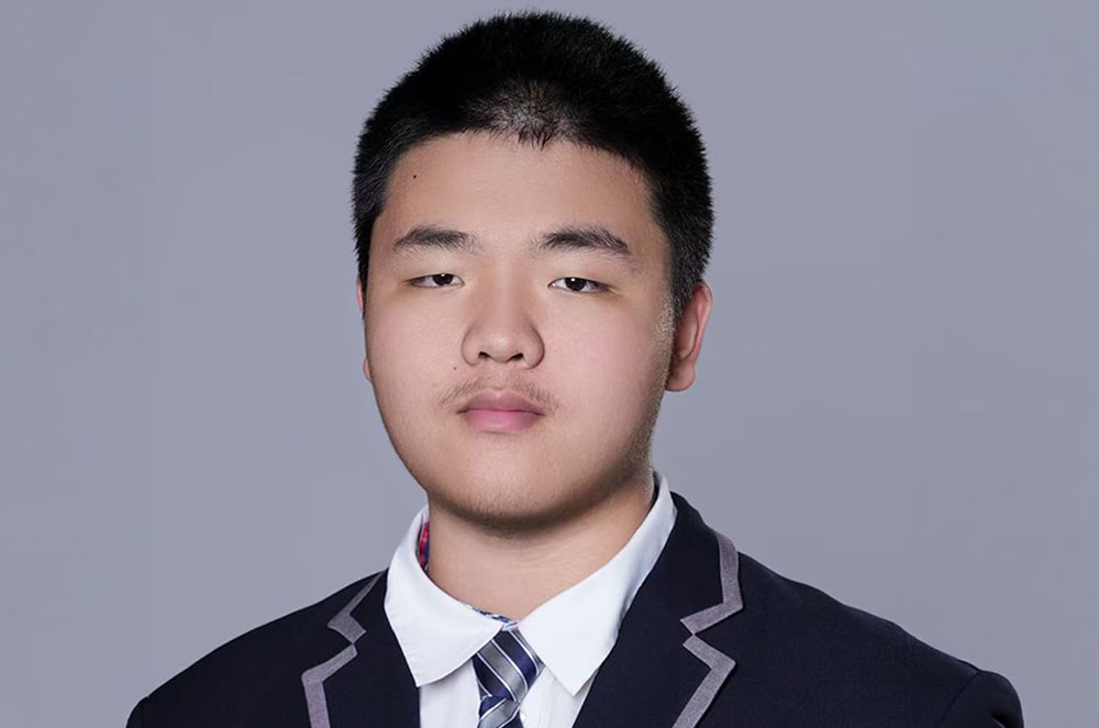 A young man in a suit and tie stands against a plain gray background.