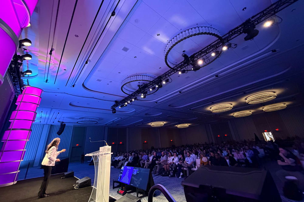 A woman stands on a stage with a podium in the foreground, addressing a large audience in a brightly lit conference hall in the background.