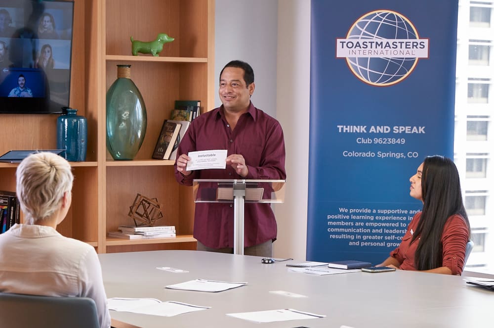 A man standing at a lectern speaking to in-person and online attendees during a meeting