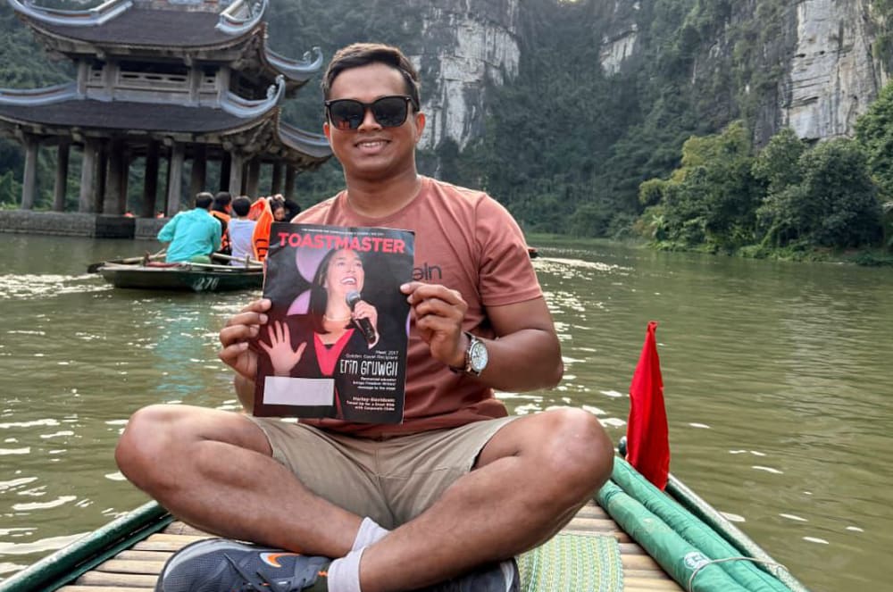 A smiling man holding a magazine and sitting on a bamboo boat