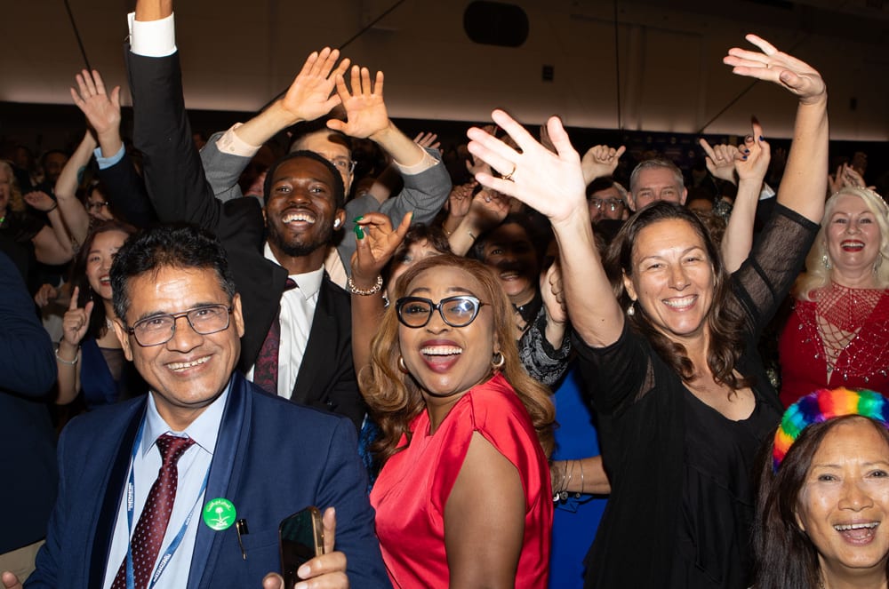 A diverse group of people are joyfully waving their hands in the foreground, with a blurred crowd visible in the background.