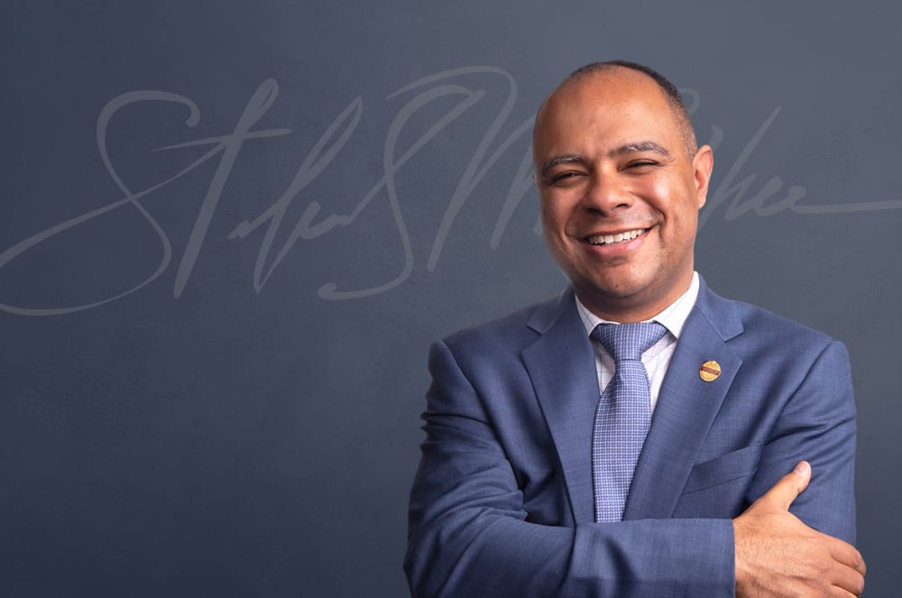 A man wearing a suit and tie smiles and crosses him arms