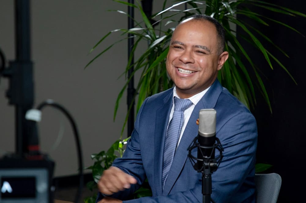A smiling man wearing a suit and tie sitting behind a microphone