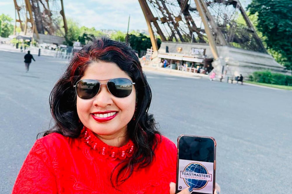 A smiling woman wearing a red shirt pointing to the Toastmasters International logo on her phone while standing near the Eiffel Tower