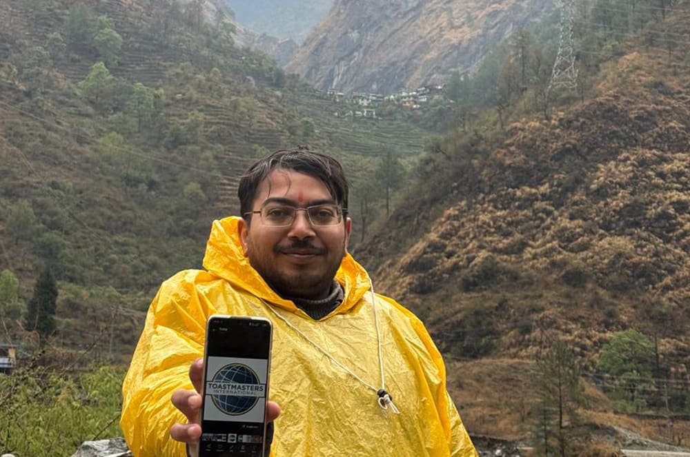 A smiling man wearing a yellow rain poncho showing the Toastmasters International logo on his phone