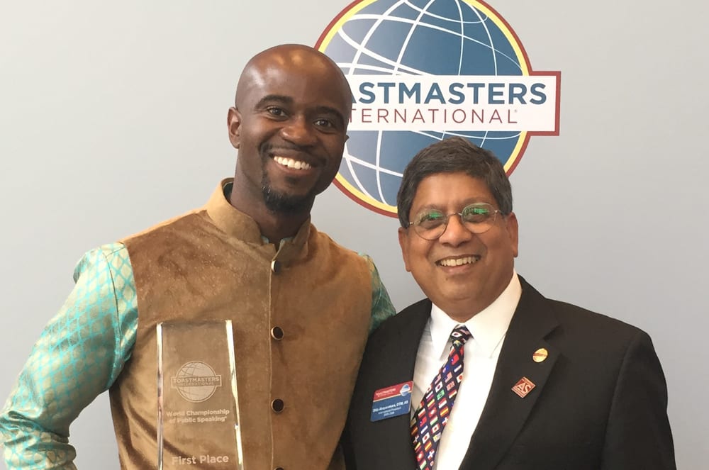 Two smiling men stand side-by-side in the foreground, with one holding a first-place trophy, against a background featuring a Toastmasters International logo.