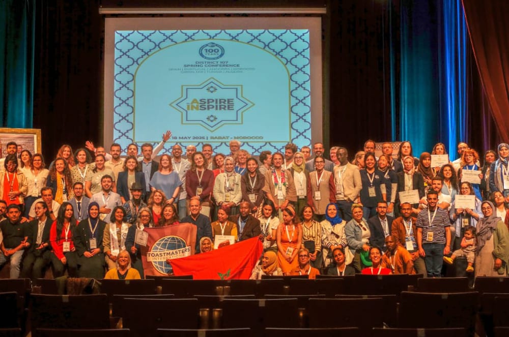 A large group of people pose for a photo in the foreground in front of a projection screen for the District 107 Spring Conference in the background.