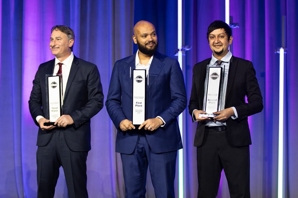Three men in suits stand side-by-side holding awards in front of a purple-lit curtain backdrop.