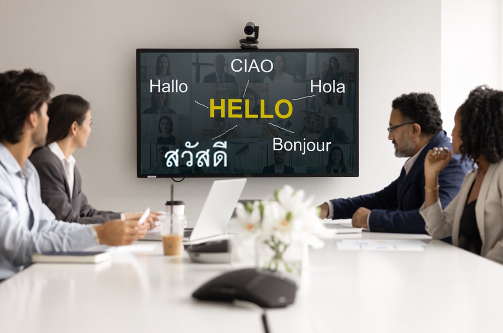 Four professionals sit around a white conference table in the foreground, facing a large screen in the background that displays "HELLO" in various languages.