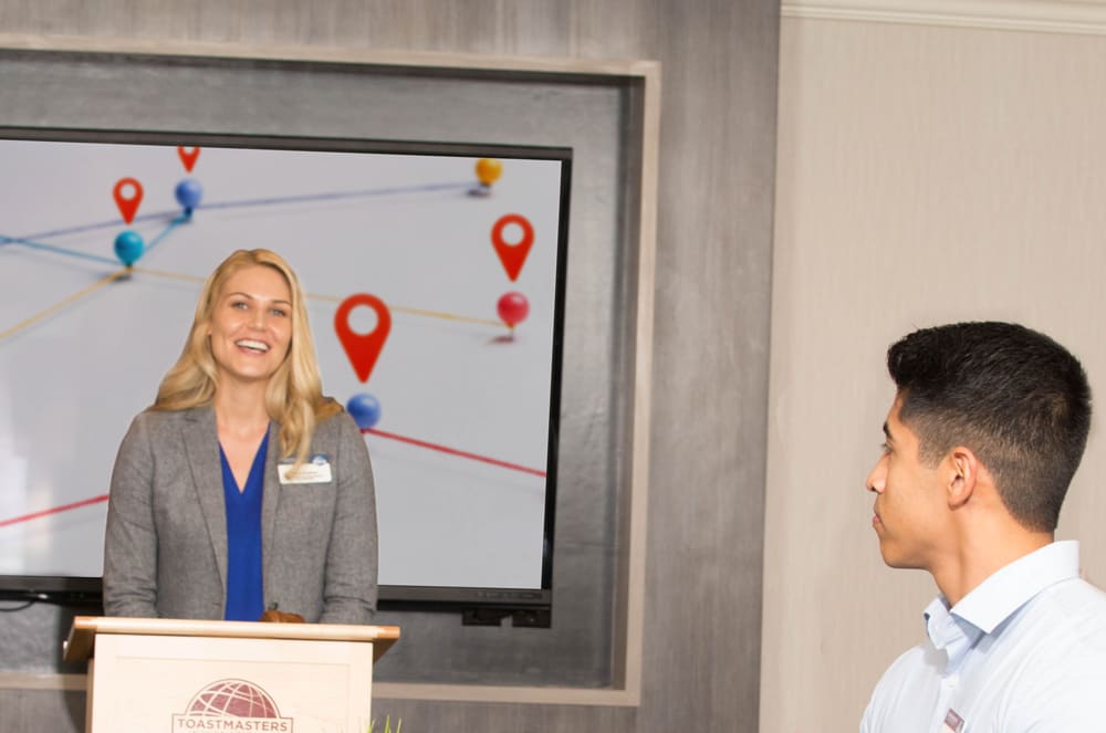 A smiling woman in a grey blazer stands at a Toastmasters podium in the foreground, while a man looks on and a screen with a map of location pins displays in the background.