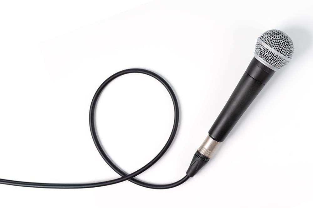 A black microphone with a silver mesh grille sits on a white background, connected to a black cable.