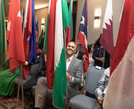 The image shows a man in a suit standing among various national flags in what appears to be a conference or meeting room.