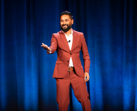 A man in a red suit stands on a stage with a blue curtain backdrop, gesturing with his hands as he speaks.