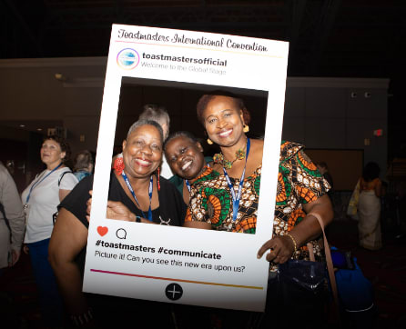 The image shows a group of three smiling women posing in front of a Toastmasters-branded photo frame, with a Toastmasters logo and hashtag visible in the background.