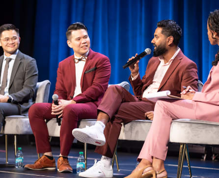 The image shows a panel discussion or event with four men sitting on a stage, dressed in stylish suits and engaging in conversation. The background is a blue curtain, creating a professional and formal setting for the discussion.