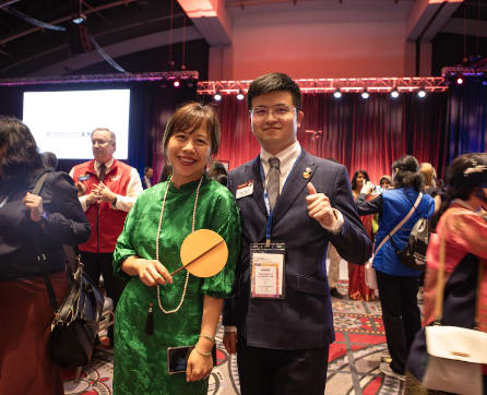 The image shows a man and a woman posing together at what appears to be a conference or event, with a colorful and lively background of people and decorations.
