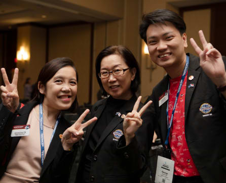 Three people, two women and one man, are posing together and making peace signs with their hands. They appear to be at some kind of event or conference, as they are wearing name tags and the background suggests a conference or meeting setting.
