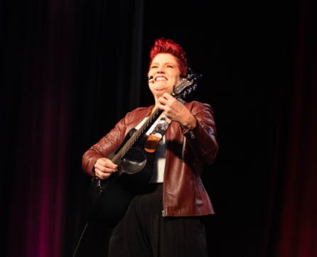 A woman with vibrant red hair is performing on stage, holding a guitar and smiling brightly against a dark background with a red curtain.
