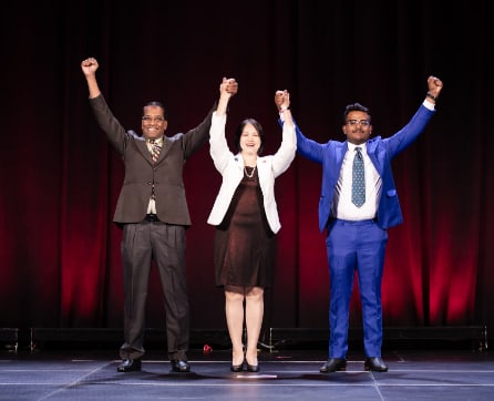 Three individuals, two men and one woman, are standing on a stage with their arms raised in a celebratory gesture against a red curtain backdrop.