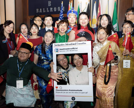 A group of people, some wearing traditional cultural attire, are gathered together and holding a sign for a Toastmasters International event.