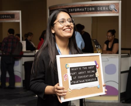 A smiling young woman holding a sign with a question about the best part of public speaking conventions, standing in front of an information desk in a crowded room.