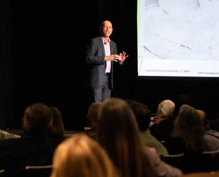 A man in a suit is standing on a stage, gesturing and speaking to an audience seated in front of him, with a large projection screen visible in the background.