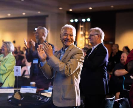 The image shows a group of people, including an older man in a light-colored suit, applauding and smiling in what appears to be a conference or event setting.