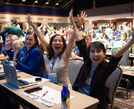 A group of enthusiastic people with their arms raised in celebration, surrounded by a lively conference or event setting.