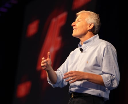 A man in a light-colored shirt stands on a stage, gesturing with his hands as he speaks to an audience in the dimly lit background.