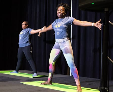 A woman in a blue shirt and colorful leggings is standing on a stage with a curtain backdrop, appearing to be engaged in some form of physical activity or performance.