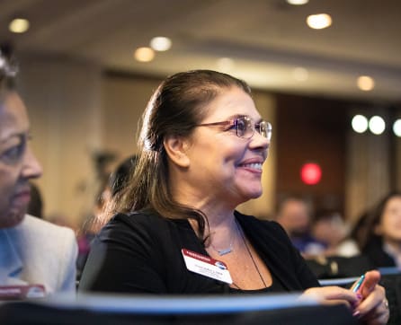 A smiling woman wearing glasses and a black jacket is seated at a table in what appears to be a conference or event setting, with other people visible in the background.