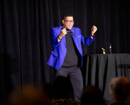 A man in a bright blue suit is standing on a stage, gesturing enthusiastically while speaking to an audience in the background.