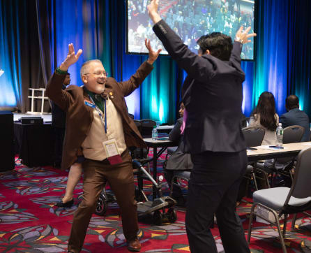 Two men on a stage, one with his arms raised in celebration, the other gesturing enthusiastically, surrounded by a colorful, patterned backdrop and an audience in the background.