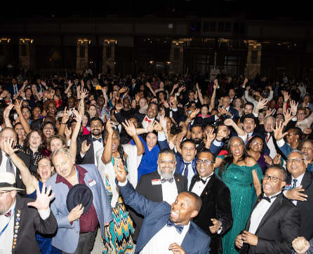 A large crowd of people, many dressed formally, are gathered together and raising their hands enthusiastically in what appears to be a celebratory event or gathering.