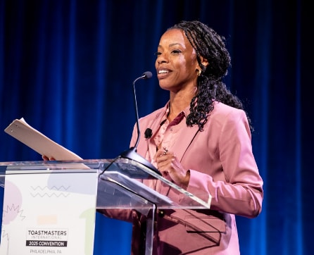 A woman with braided hair is standing at a podium, speaking into a microphone while wearing a pink blazer against a blue curtain backdrop.