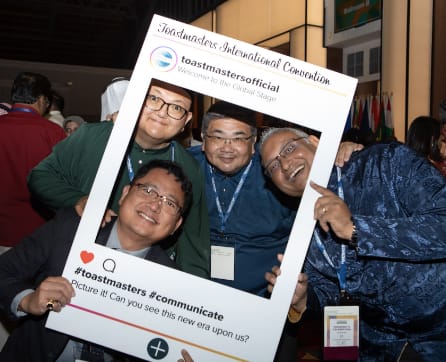 The image shows a group of people posing together in front of a large frame that appears to be a promotional display for Toastmasters, an organization focused on communication and leadership development.