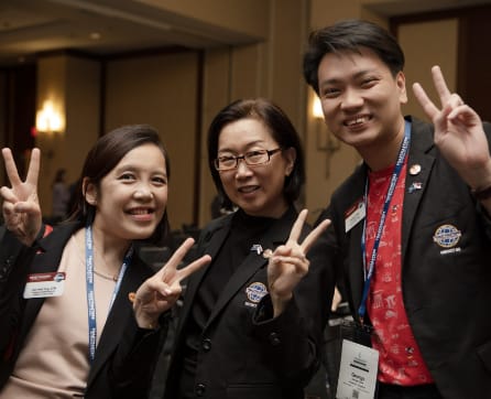 Three people, two women and one man, are standing together and making peace signs with their hands. They appear to be at some kind of event or conference, as they are wearing name tags and the background suggests a conference or meeting setting.