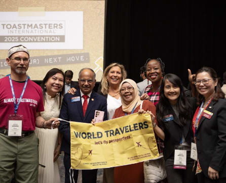 A group of people, some wearing Toastmasters convention attire, are gathered together holding a 'World Travelers' banner in the foreground, with a Toastmasters convention backdrop visible in the background.