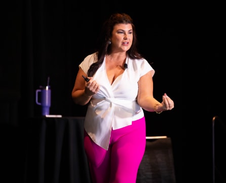 A woman in a white blouse and bright pink pants stands on a stage, gesturing with her hands.